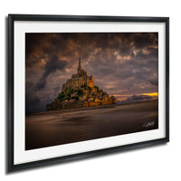 A framed photograph depicting the iconic Mont Saint-Michel, a medieval fortified island in Normandy, France, with its distinctive spire and castle-like architecture set against a dramatic sky.