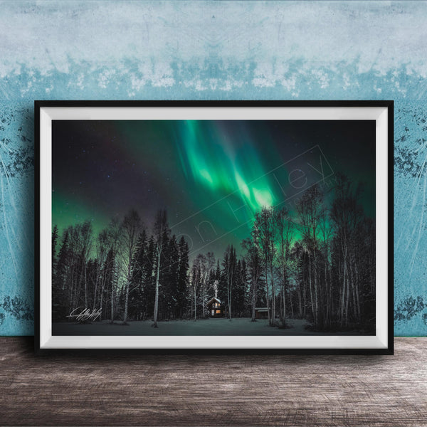A framed photograph depicting a serene night scene with a forest and a house illuminated by the vibrant green and blue hues of the Aurora Borealis ( Aurora lights) in the sky.