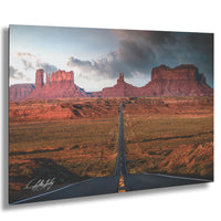 a long, straight road cutting through a vast, arid landscape with towering red rock formations in the background.