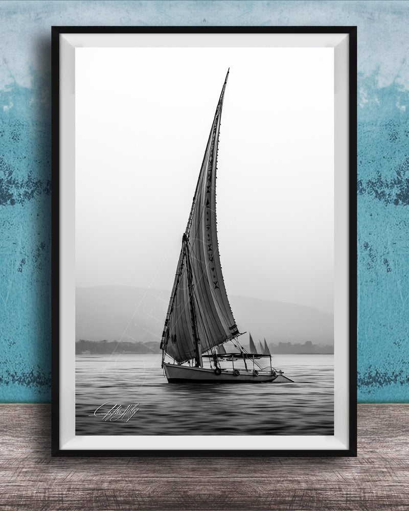 This luxury black and white fine art photograph captures a felucca sail boat drifting along the Nile River at sunset near Luxor, Egypt. Soft fog and minimalist composition create a timeless, monochrome aesthetic rooted in Egyptian river culture. Ideal for collectors and interior designers seeking refined, museum-quality wall art that embodies elegance, simplicity, and enduring calm.
