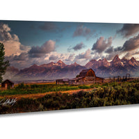 A picturesque rural landscape with a wooden barn, a small shed, and a dirt road leading to a mountain range in the distance. The sky is filled with clouds, and the sun is setting, casting a warm glow over the scene.