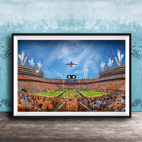 A framed photograph depicting a large crowd of people gathered in a stadium, with a plane flying overhead and fireworks lighting up the sky.