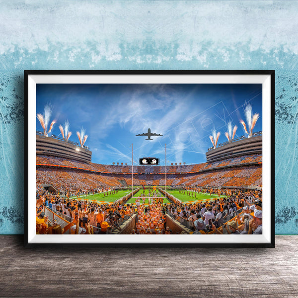 A framed photograph depicting a large crowd of people gathered in a stadium, with a plane flying overhead and fireworks lighting up the sky.
