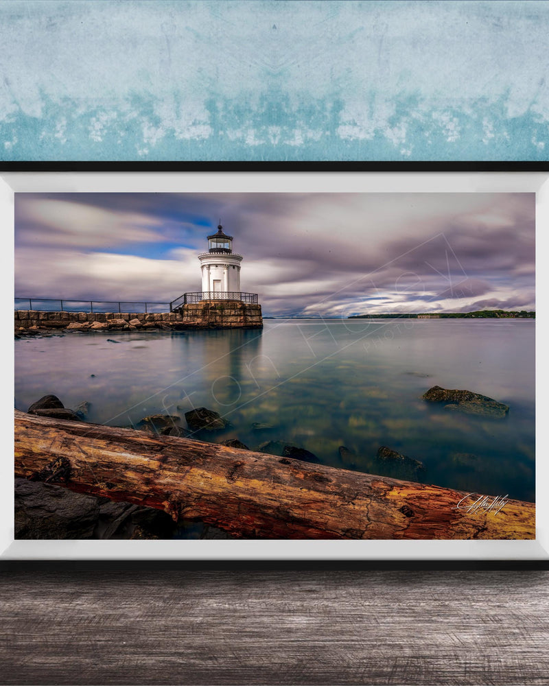 A framed photograph of a lighthouse on a rocky shore, with a calm body of water in the background and a cloudy sky above.