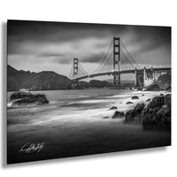 A black and white photograph of the Golden Gate Bridge in San Francisco, California, with the ocean and rocky coastline in the foreground.