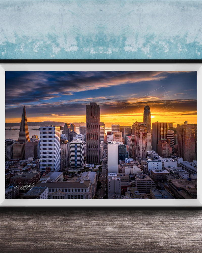 The image is a framed photograph of a city skyline at sunset, with the sun setting behind the buildings and casting a warm, golden glow over the urban landscape.