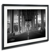 A black and white photograph of a bridge with tall columns and arches, leading to a road or walkway. The bridge appears to be an architectural landmark, with the surrounding buildings and trees visible in the background.
