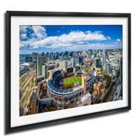An aerial view of a large city with a baseball stadium in the center, surrounded by tall buildings and a clear blue sky with white clouds.