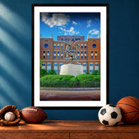 A framed photograph of a statue of a person on a pedestal in front of a brick building, with a basketball, soccer ball, and baseball glove on a wooden surface below the frame.