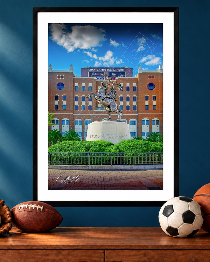 A framed photograph of a statue of a person on a pedestal in front of a brick building, with a basketball, soccer ball, and baseball glove on a wooden surface below the frame.