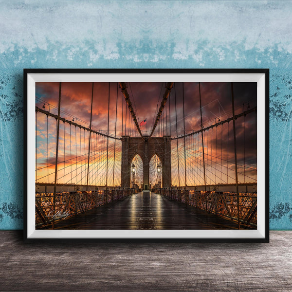 A framed photograph of the Brooklyn Bridge at sunset, with the bridge's intricate metal structure and suspension cables silhouetted against a vibrant sky filled with clouds and warm hues.