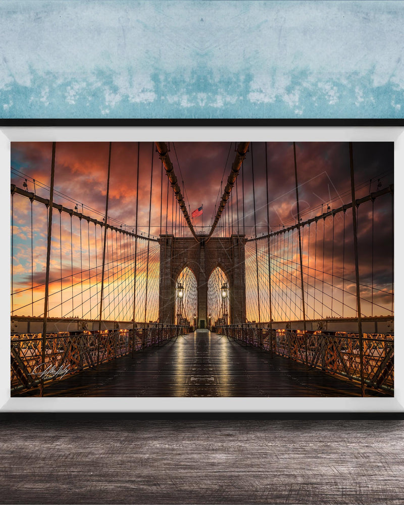 A framed photograph of the Brooklyn Bridge at sunset, with the bridge's intricate metal structure and suspension cables silhouetted against a vibrant sky filled with clouds and warm hues.