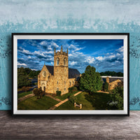 A stone church with a tall tower, surrounded by trees and a grassy area, set against a blue sky with clouds.