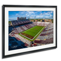 The image is a framed photograph of a football stadium with a large field, surrounded by rows of seating and a scoreboard.