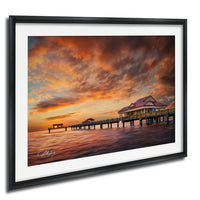 A framed photograph depicting a serene sunset over a wooden pier with a small building on it, set against a vibrant sky filled with clouds.