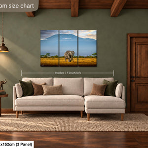 The image is a livingroom with a large painting of an elephant in a field hanging on the wall. The painting is divided into three panels, and the livingroom has a beige sofa and a wooden door.