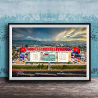 A framed photograph of a large sports stadium with a red roof, surrounded by mountains and a cloudy sky.