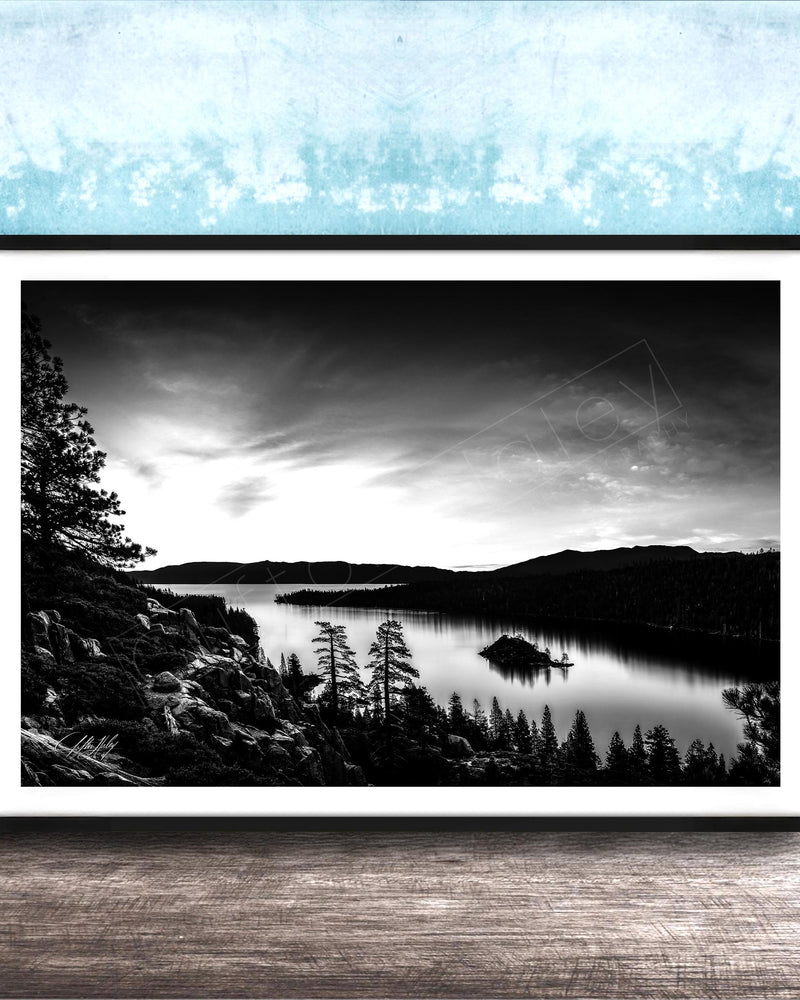A black and white landscape photograph depicting a serene lake surrounded by mountains and pine trees, with a cloudy sky overhead.