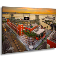 The image is an aerial view of a large stadium with a red and white exterior, surrounded by palm trees and other buildings. The stadium appears to be a sports venue, with a large scoreboard visible in the background. The sky is filled with warm, orange and yellow hues, suggesting a sunset or sunrise. The image is a collage of multiple images, and the artist's signature is visible in the bottom left corner.