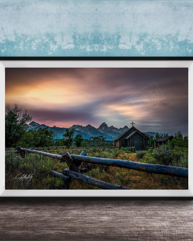 A framed photograph depicting a serene rural landscape with a wooden fence, a church, and mountains in the distance, set against a colorful sunset sky.