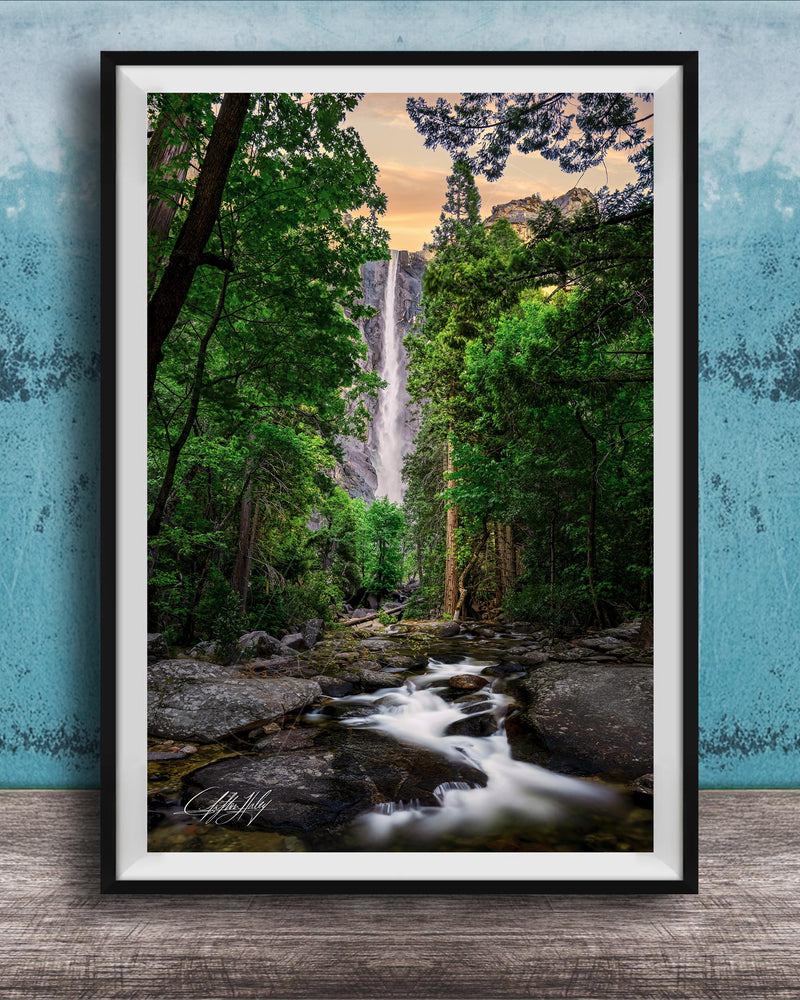 A framed photograph of a waterfall cascading through a lush, forested landscape, with the water flowing over rocks and surrounded by trees.