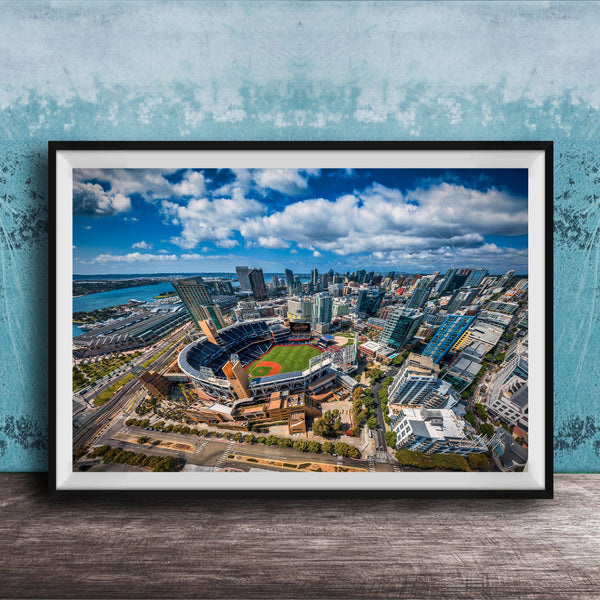 The image is an aerial view of a city with a baseball stadium in the center, surrounded by tall buildings and a body of water.