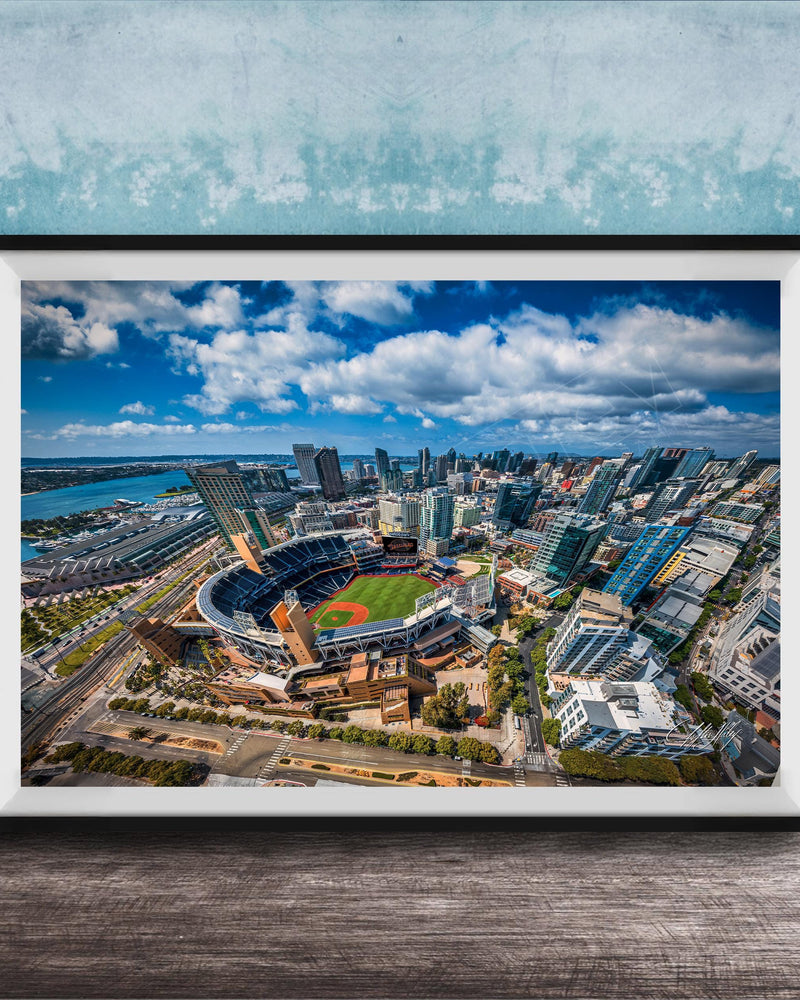 The image is an aerial view of a city with a baseball stadium in the center, surrounded by tall buildings and a body of water.