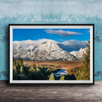 A framed photograph of a mountainous landscape with snow-capped peaks, a river, and a forested area in the foreground.