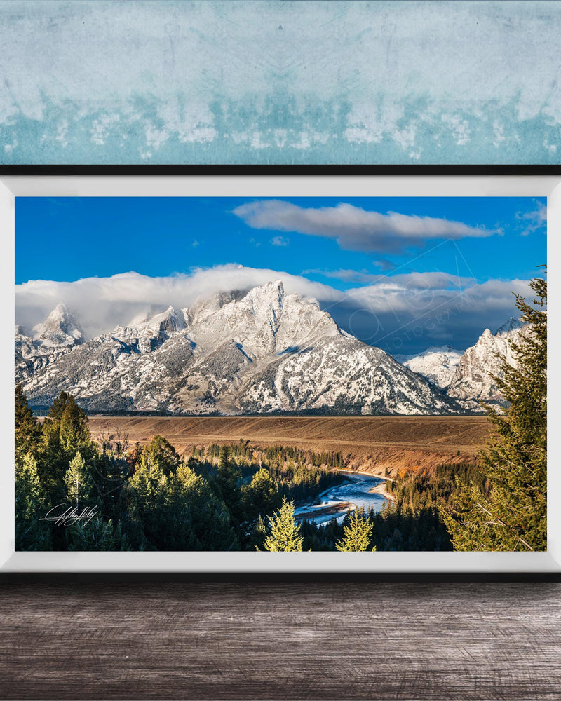 A framed photograph of a mountainous landscape with snow-capped peaks, a river, and a forested area in the foreground.
