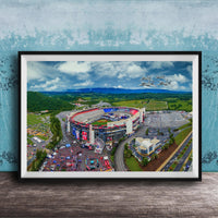 The image is an aerial view of a large stadium surrounded by a green landscape, with a few buildings and vehicles visible.
