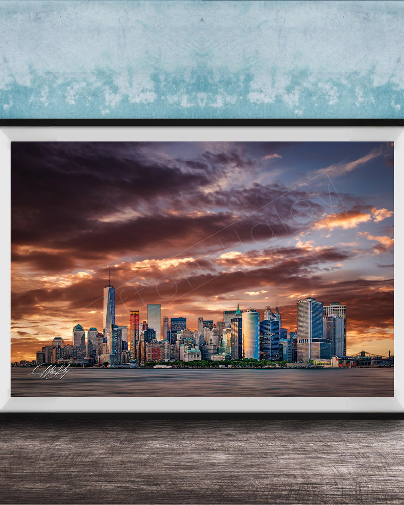 A framed photograph of the New York City skyline at sunset, with the city's skyscrapers silhouetted against a dramatic sky filled with clouds and vibrant colors.