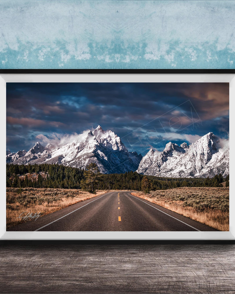 A framed photograph of a winding road leading towards majestic snow-capped mountains against a dramatic cloudy sky.