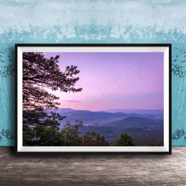 A framed photograph of a scenic mountain landscape at sunset, with a silhouetted tree in the foreground and a hazy, purple sky in the background.
