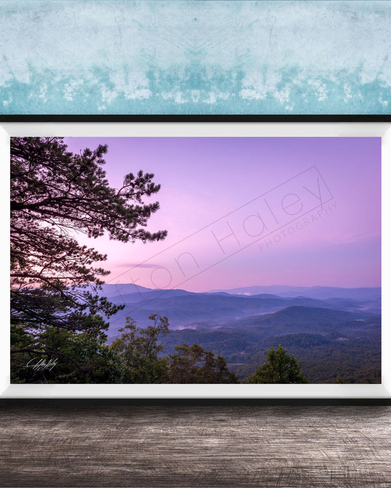 A framed photograph of a scenic mountain landscape at sunset, with a silhouetted tree in the foreground and a hazy, purple sky in the background.