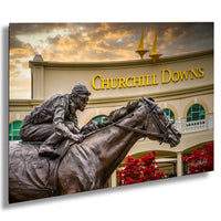 May include: A bronze statue of a jockey riding a horse, the statue is in front of a building with the words 'CHURCHILL DOWNS' in gold letters. The statue is in front of a building with a white facade and red bushes in front of it.