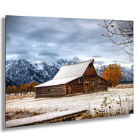 A rustic wooden barn stands in a snowy field, with snow-capped mountains in the background and a few trees with orange foliage.
