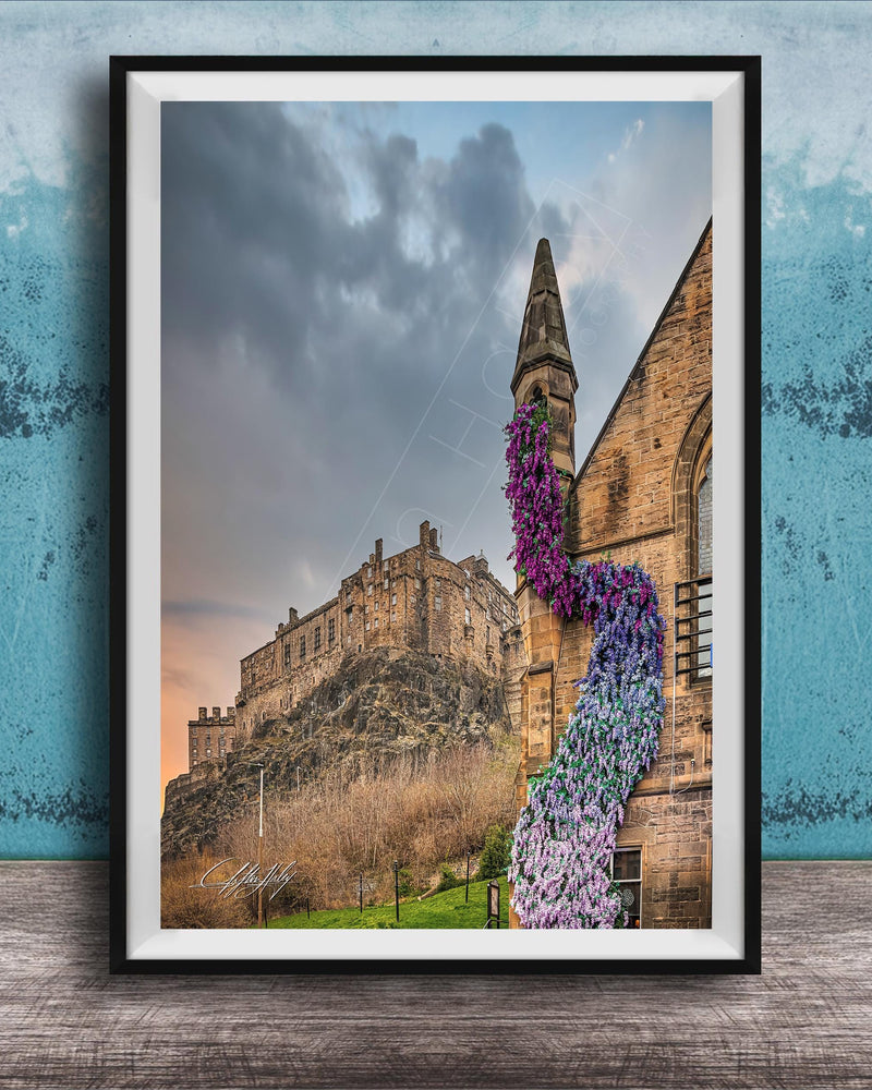 A framed photograph of a castle with a tower, set against a cloudy sky, with a colorful flower arrangement hanging from the tower.