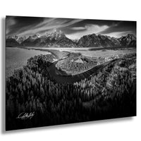 A black and white landscape photograph depicting a winding river surrounded by a forested area with snow-capped mountains in the background.