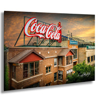 A large Coca-Cola sign is displayed on top of a brick building, with a stadium and other buildings visible in the background.